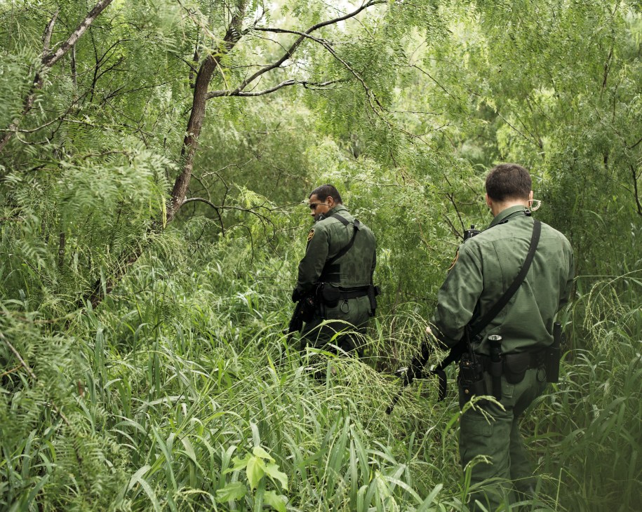 Two US Border Patrol agents in green uniforms walk through dense vegetation at a border crossing point in Texas.