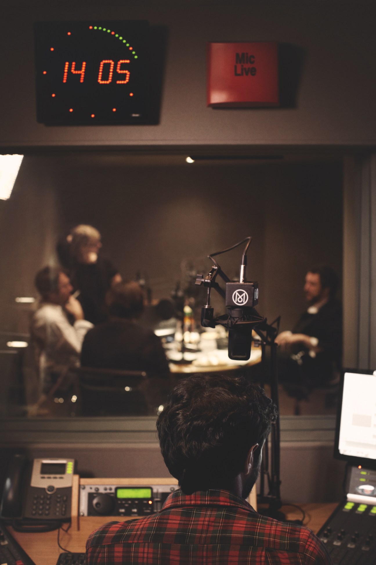 Engineer at radio studio desk with microphone and equipment, monitoring broadcast with studio visible through window.