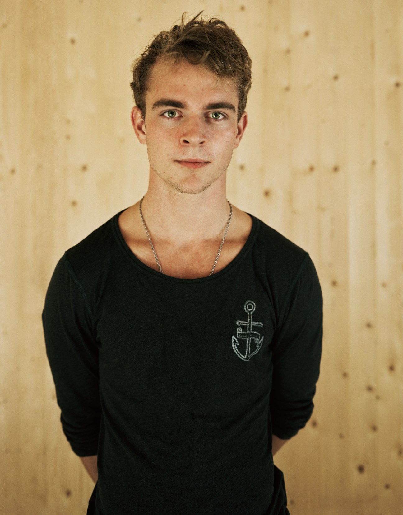 Young man wearing a black long-sleeved shirt with an anchor design against a textured beige background.