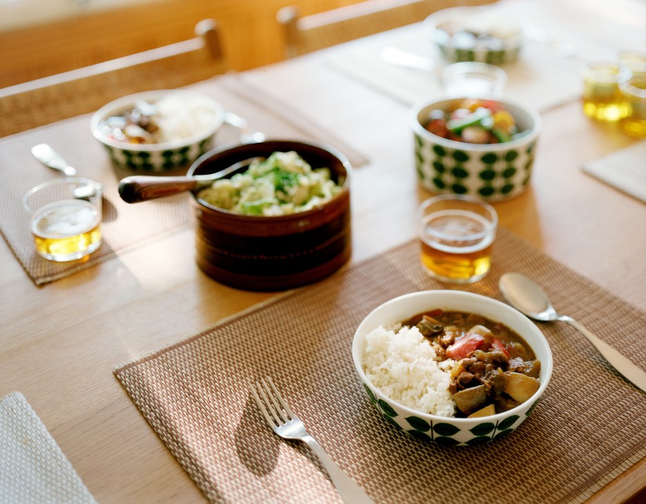 Japanese curry served in patterned bowls with rice on a wooden dining table with beer and place settings.