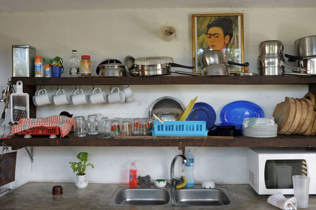 Kitchen shelves with cookware, dishes, and a Frida Kahlo portrait above a double sink.