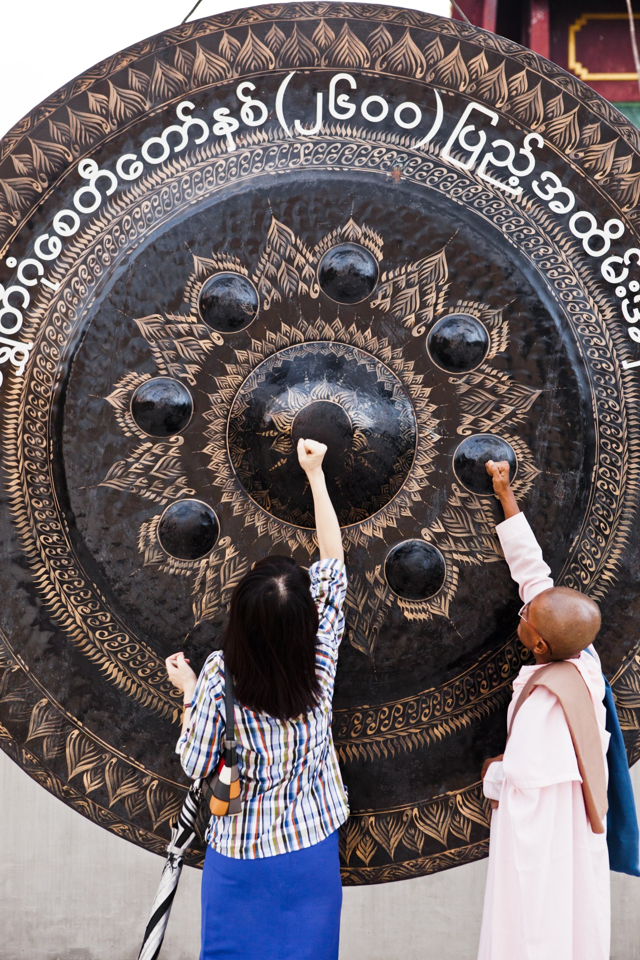 Two people striking a large ornate bronze gong with intricate gold leaf patterns at Shwedagon pagoda.