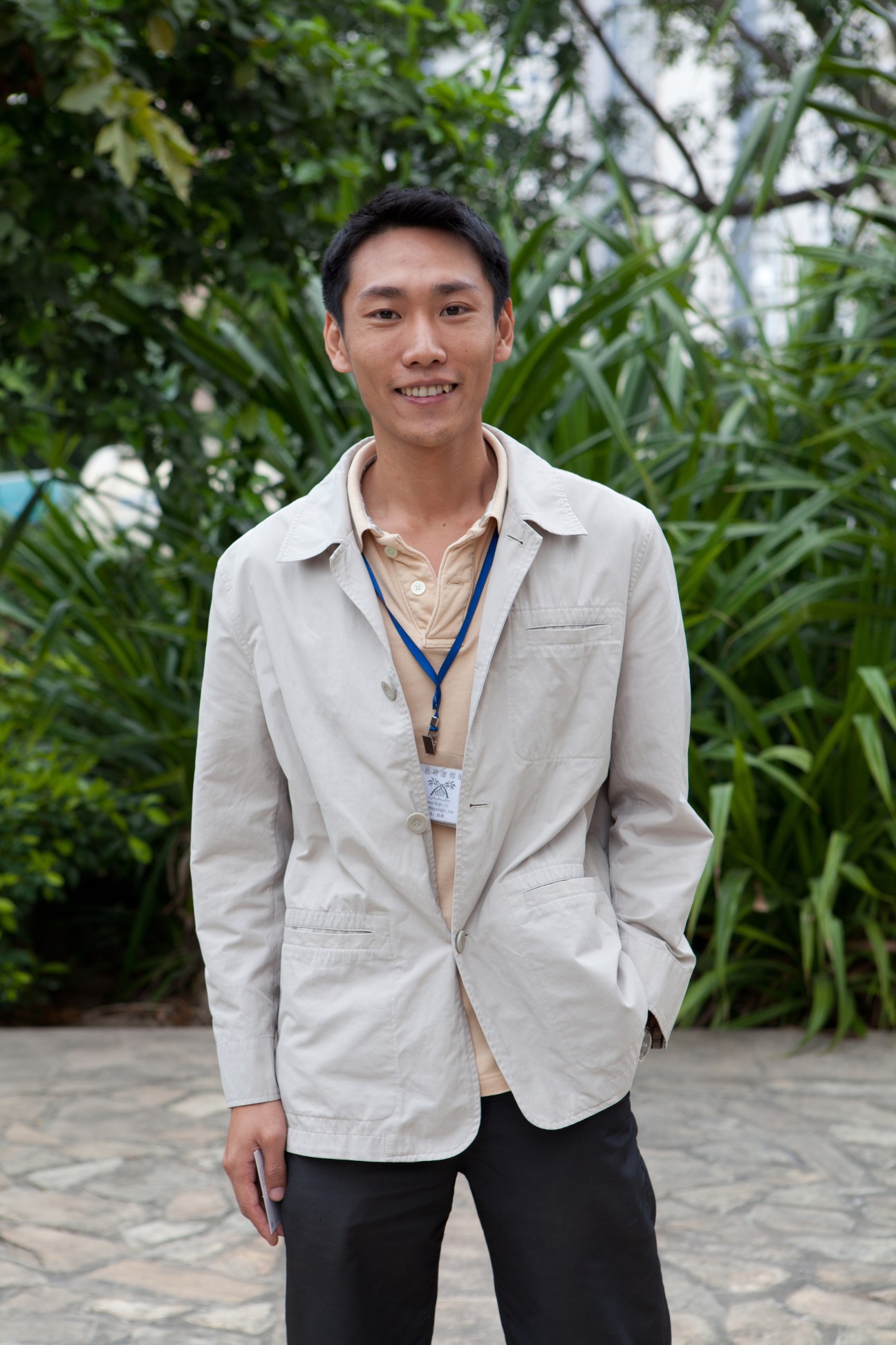 Alan Lin, serviced apartments manager at BaGe Holiday Inn, wearing a light beige blazer and ID badge, smiling in front of tropical plants.