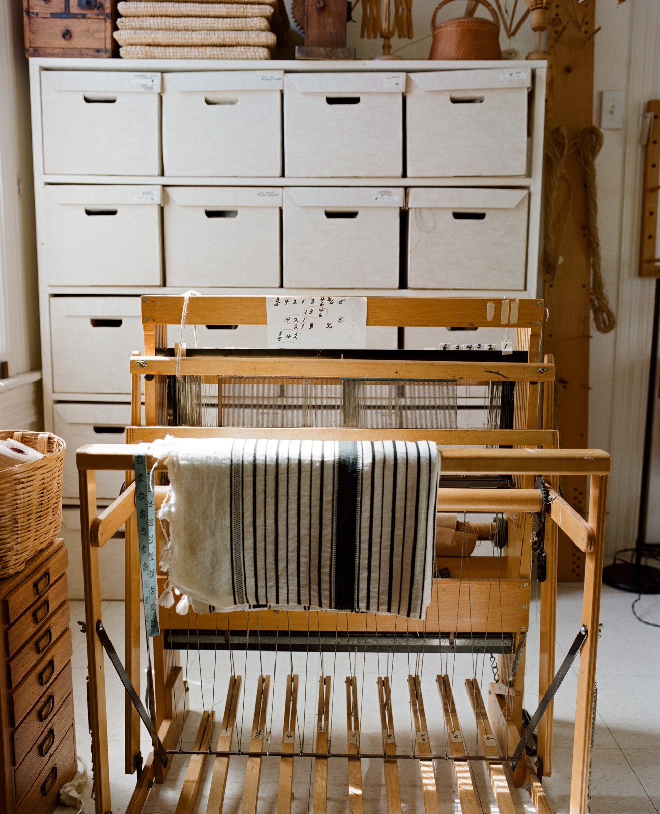 A wooden loom with striped fabric woven across it, with a white storage unit containing boxes in the background.