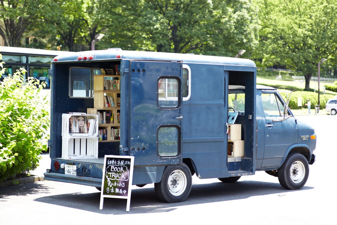 Blue vintage Chevy van converted into a mobile bookshop with wooden shelves displaying books and magazines visible through open doors.