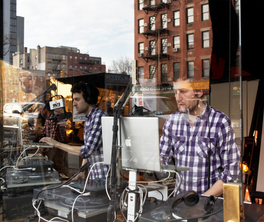 Two men in plaid shirts broadcast from a glass-fronted radio studio on a New York street with buildings visible in the background.