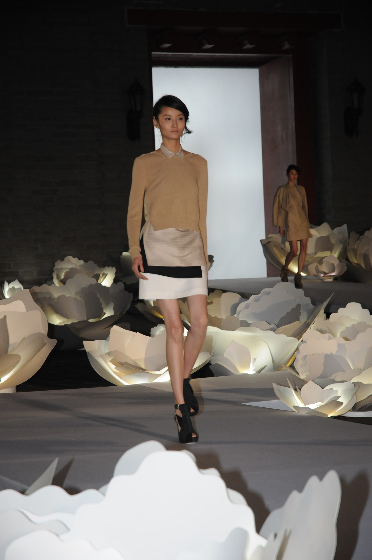 Model on catwalk wearing a beige and cream dress with black platform heels, walking past large white sculptural elements.