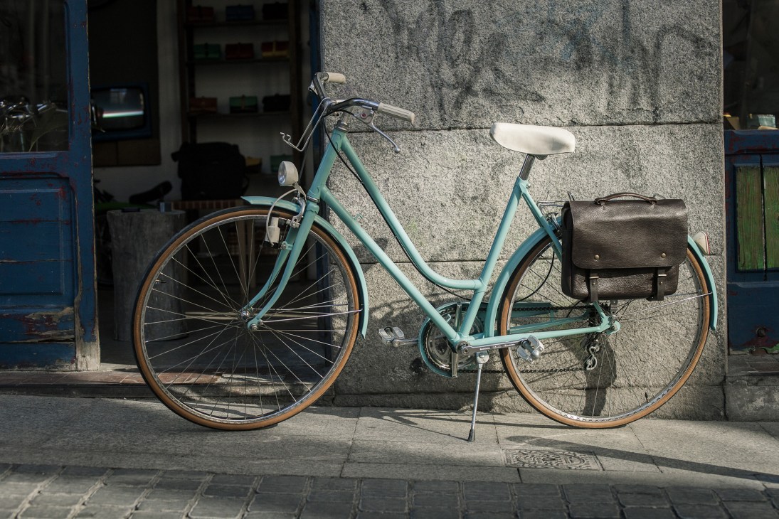 Turquoise vintage bicycle with white saddle and black leather bag leaning against concrete wall.