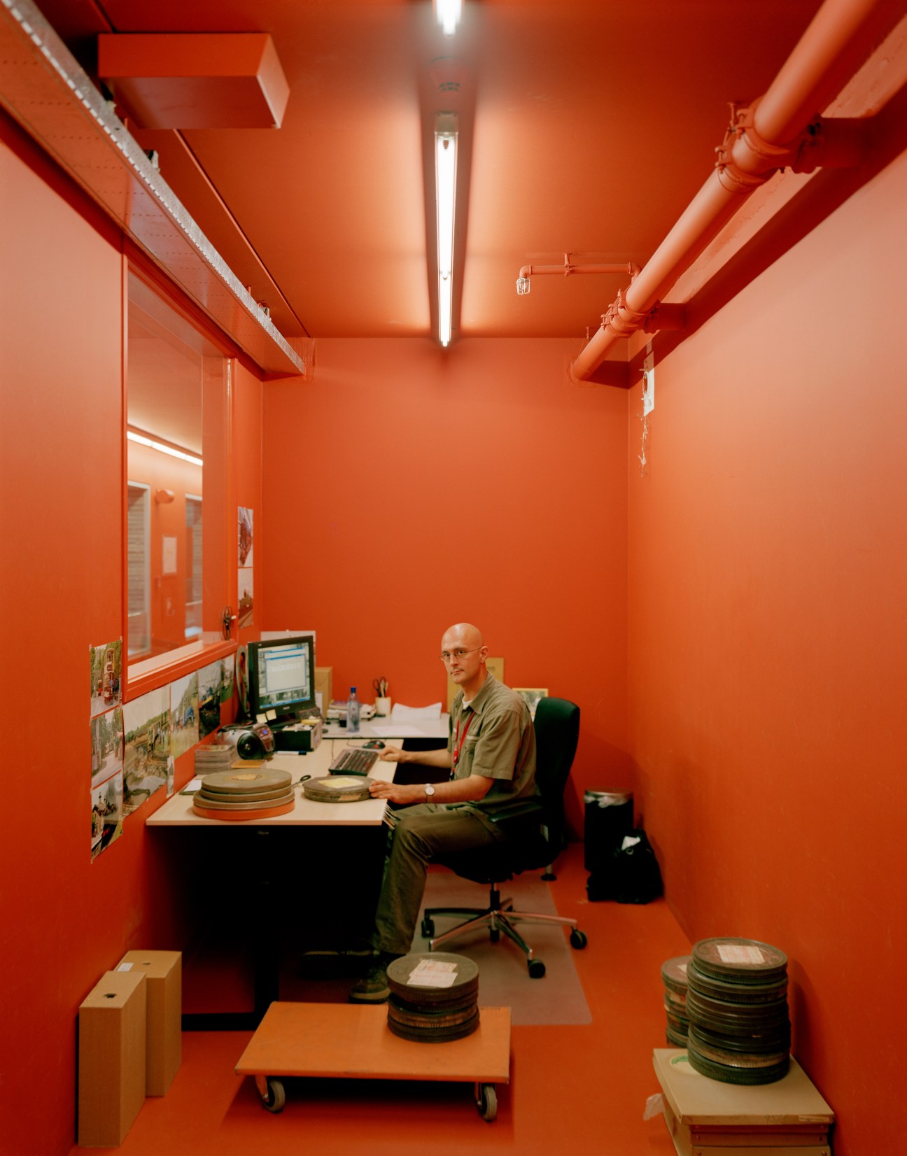 Archivist working at desk in bright orange-walled office surrounded by film reels and archival materials.