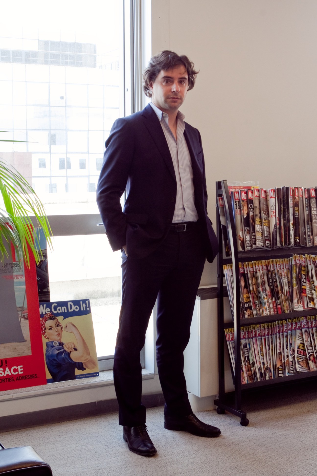 Publisher Edouard Minc standing in an office with magazines displayed on shelves behind him.
