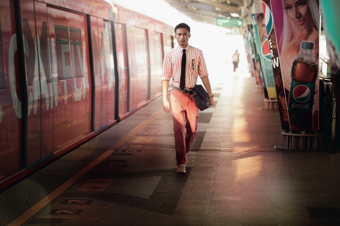 Man in pink striped shirt and coral trousers walking through Bangkok BTS Skytrain station with black bag.