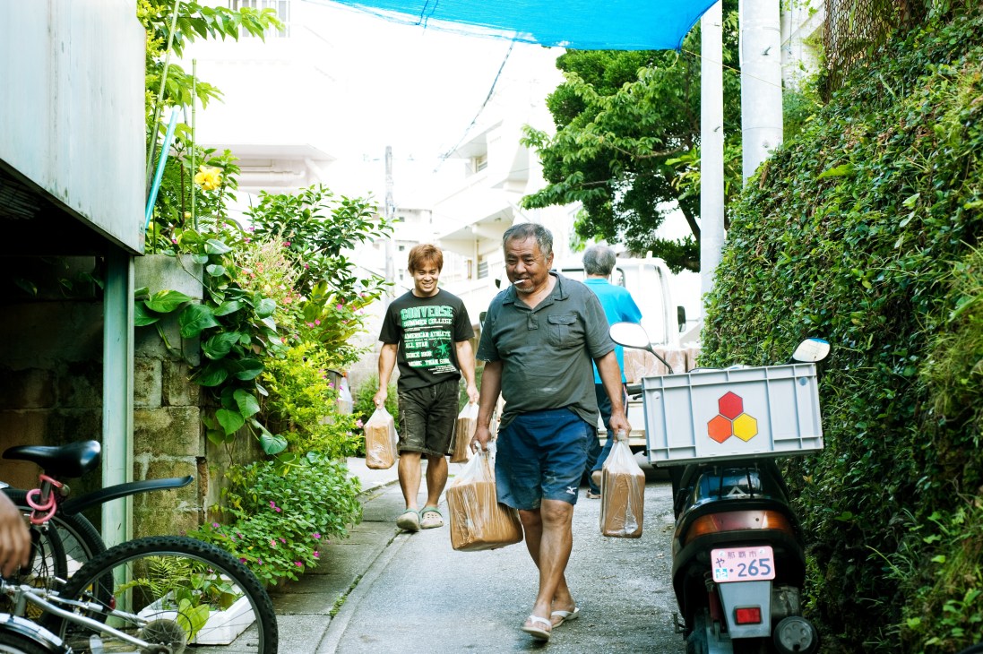 Man and boy carrying wooden containers walk down a narrow alleyway lined with lush green foliage and white buildings in Naha.
