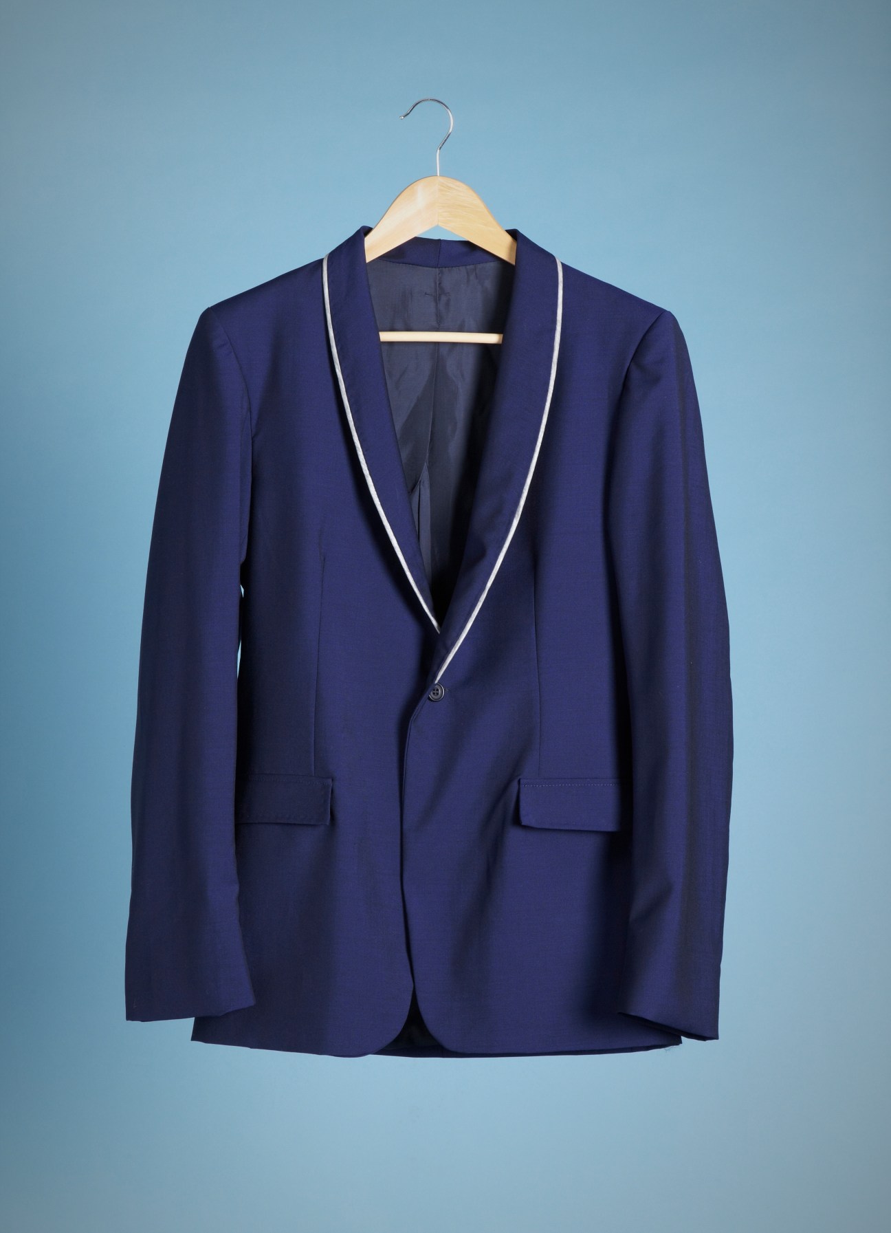 Navy blazer with white piping on wooden hanger against blue background.