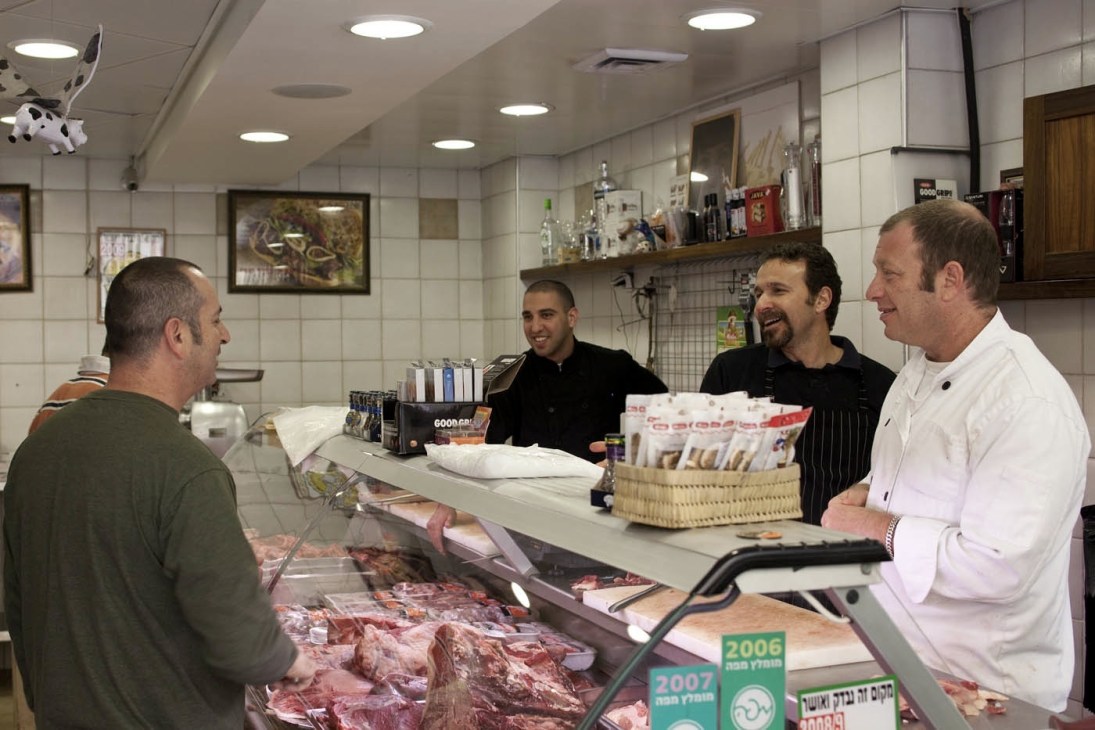 Local men gather at a butcher counter displaying fresh cuts of meat at Zalman's Meats in Tel Aviv's Noga district.