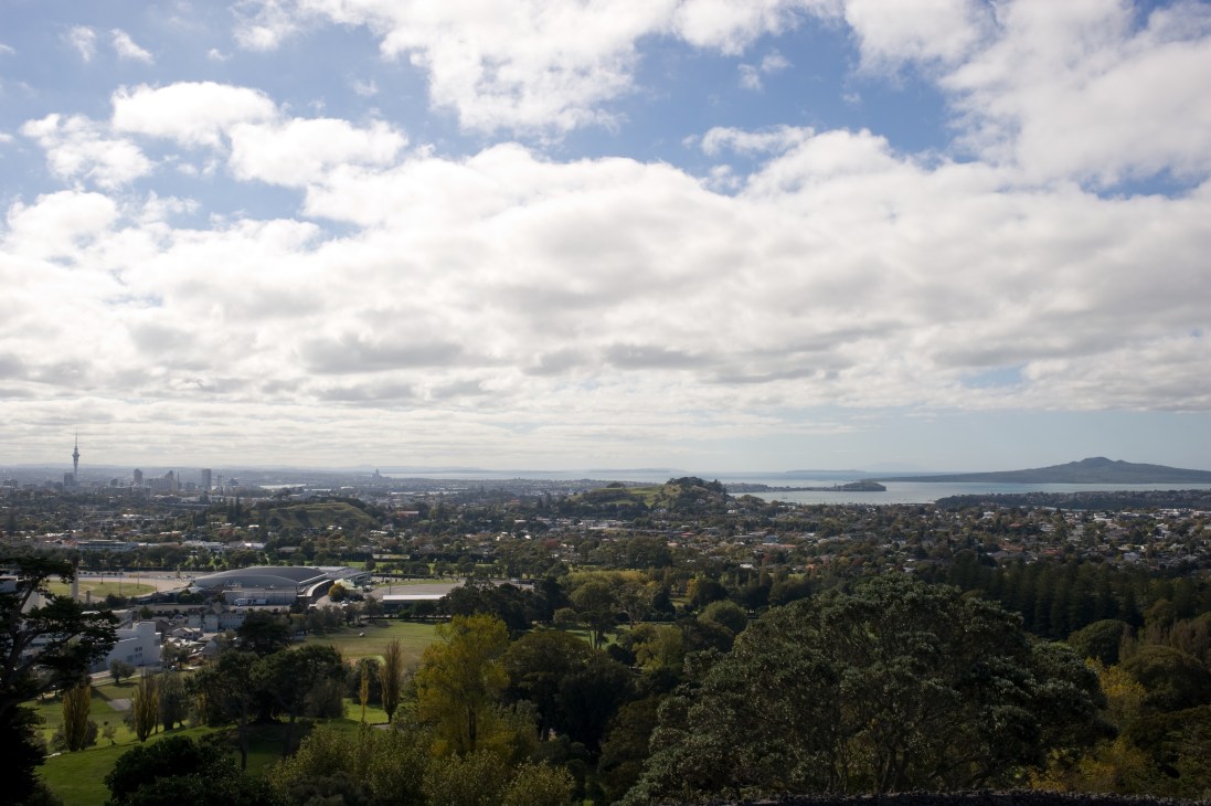 Aerial view of Auckland cityscape with central business district, green suburbs, and harbor in background under cloudy sky.