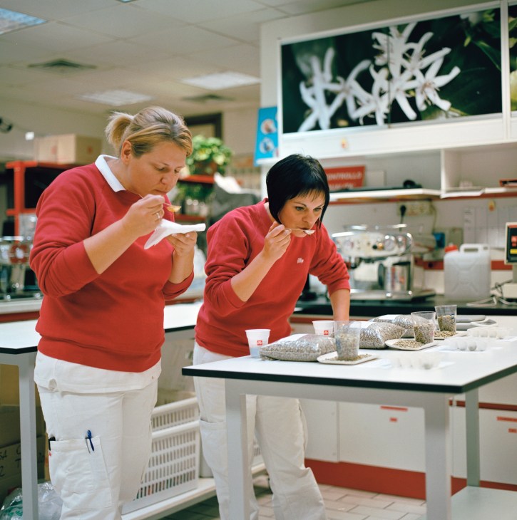 Two Illy coffee sommeliers in red shirts taste and evaluate espresso samples in a laboratory setting.