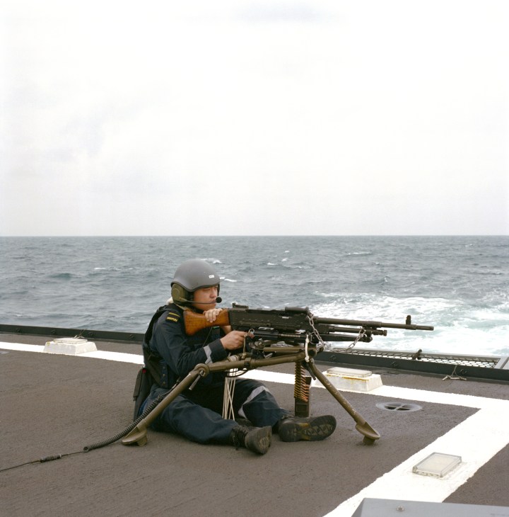 A sailor manning a machine gun on the helicopter deck of a naval frigate overlooking the sea.