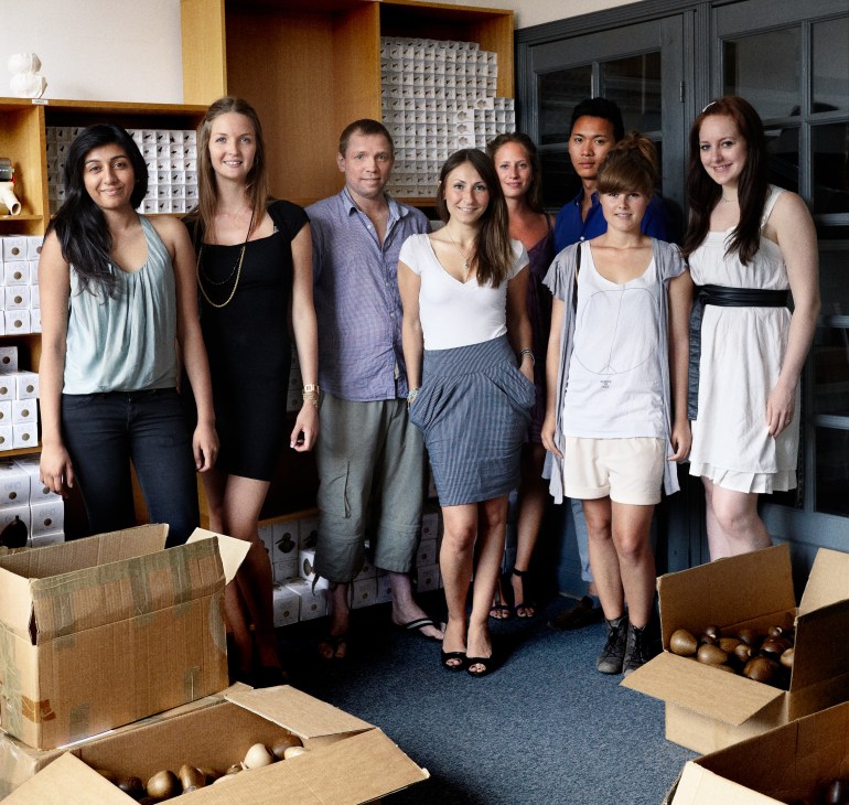 Eight young professionals pose together in a modern office space with wooden shelving and cardboard boxes containing design products.
