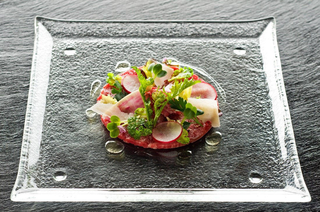 A plated dish with sliced red beef, radish, and fresh herbs on a textured slate plate.