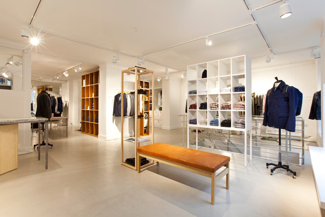 Interior of Tres Bien menswear store in Stockholm with white walls, wooden shelving, clothing racks, and an orange bench.