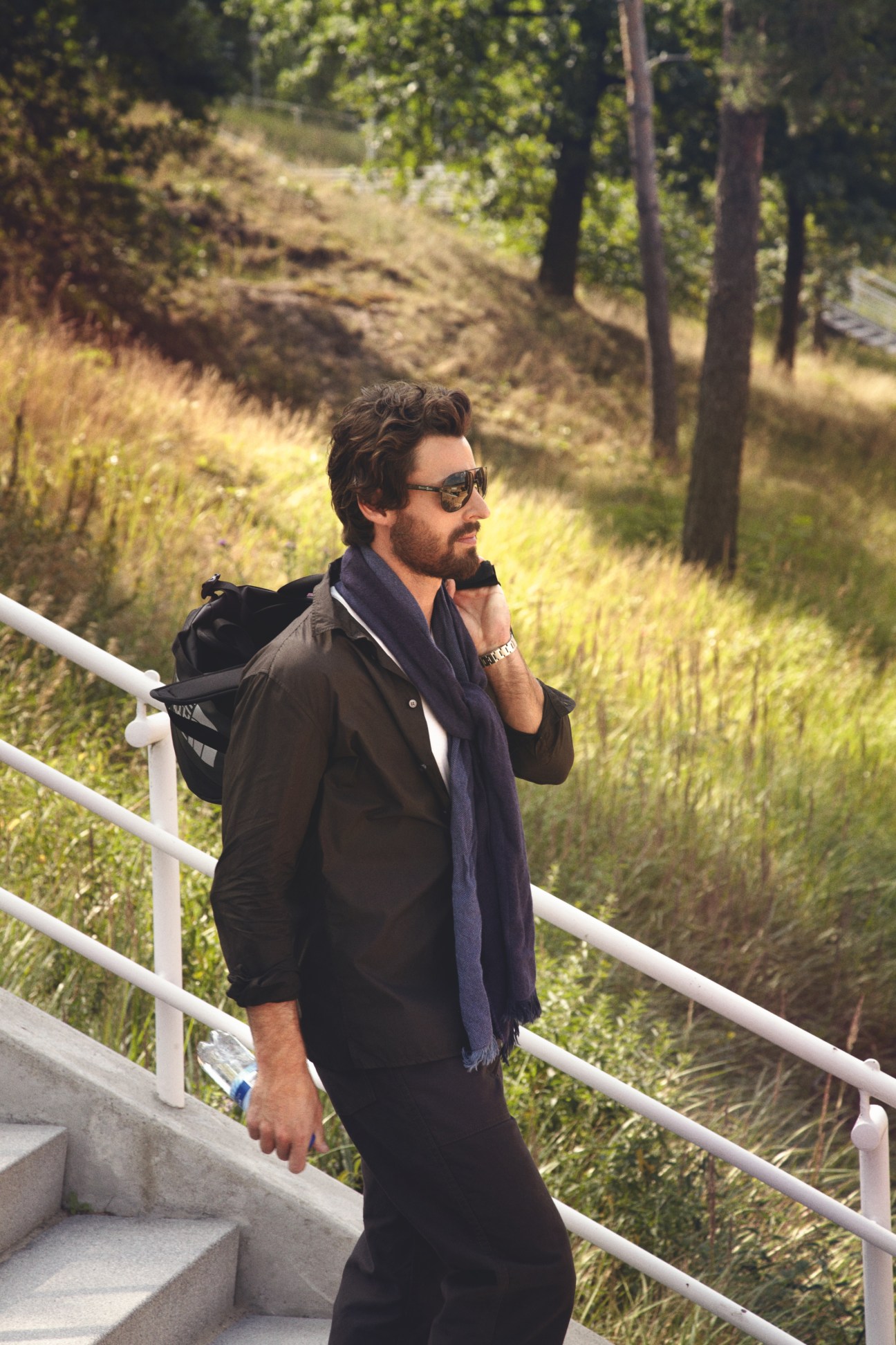 Man in Bottega Veneta shirt and White Mountaineering trousers standing on stairs overlooking forested landscape.