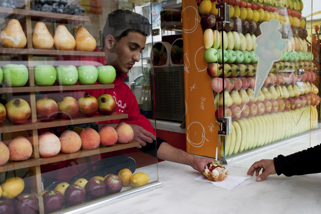 A waiter in a red shirt serves ice cream at a fruit stand displaying colorful apples and pears in Baghdad.