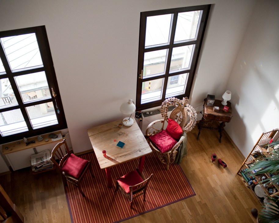 Overhead view of a bright apartment living room with red cushioned chairs, wooden desk, large windows, and hardwood flooring.