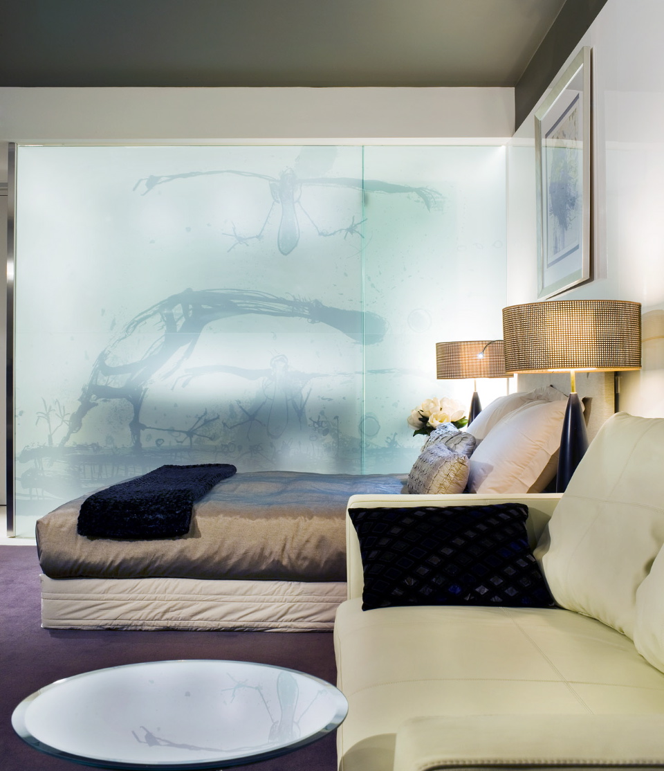 Modern hotel bedroom with bed, cream armchair, brass lamp, and large glass wall featuring John Olsen artwork.
