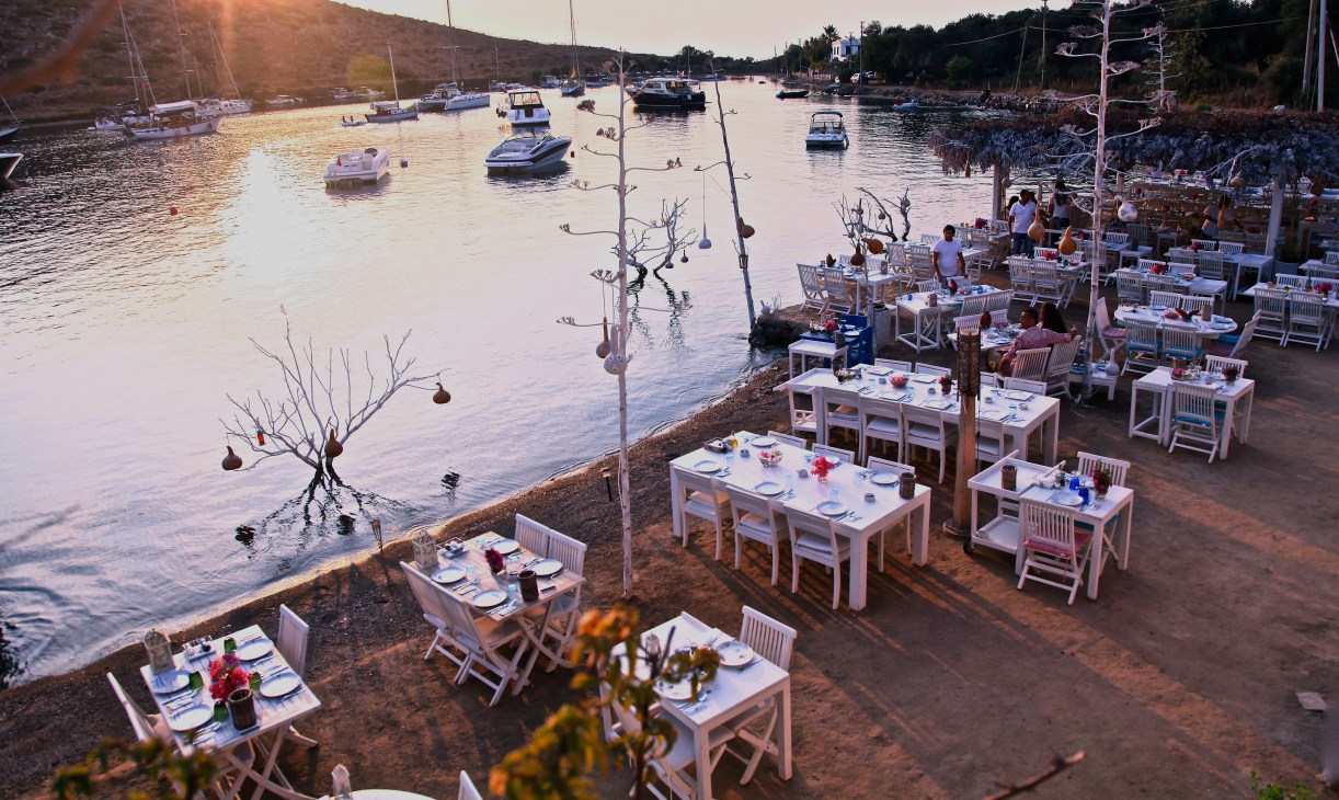 Waterfront dining setup at Mimoza restaurant with white tables and chairs along a cove at sunset, boats moored in the water beyond.