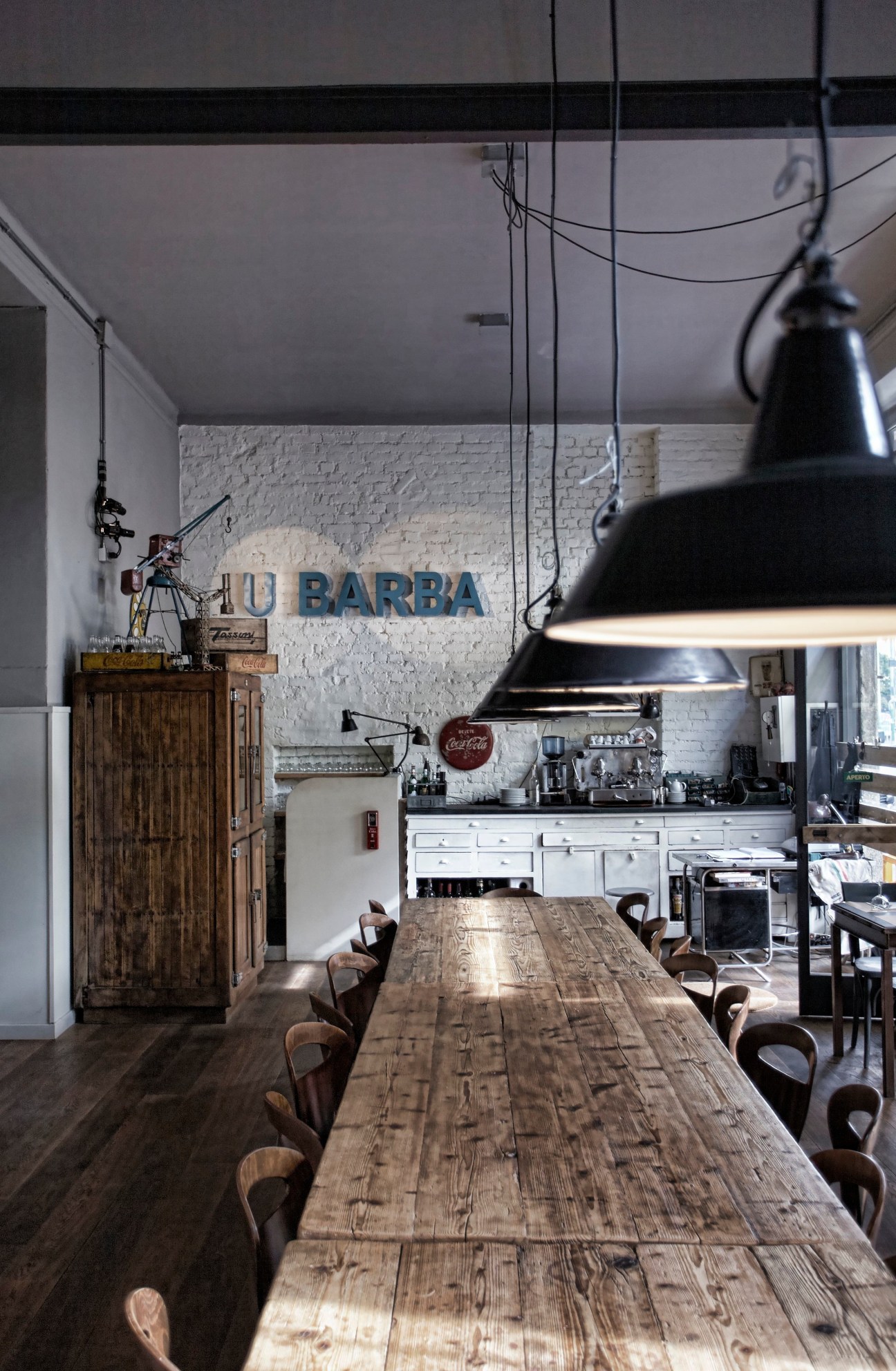 U Barba osteria in Milan with white brick walls, wooden table, industrial pendant lights, and blue signage.