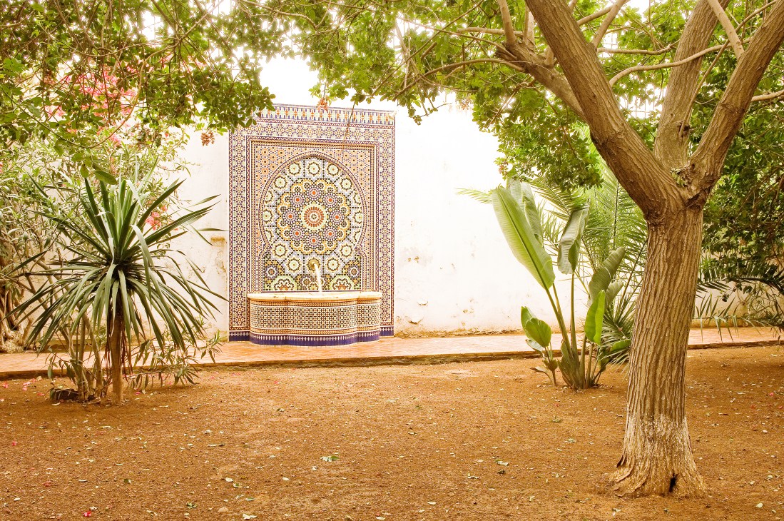 Ornate Moroccan mosaic fountain in courtyard with trees and traditional architecture.