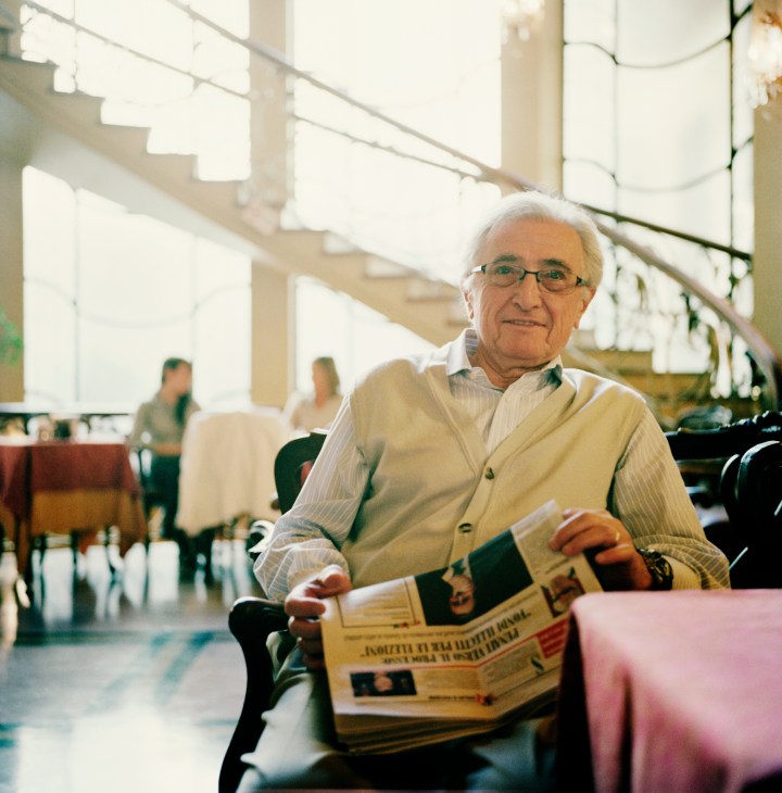 An elderly man with white hair and glasses reads a newspaper while seated at Cafe Torino.