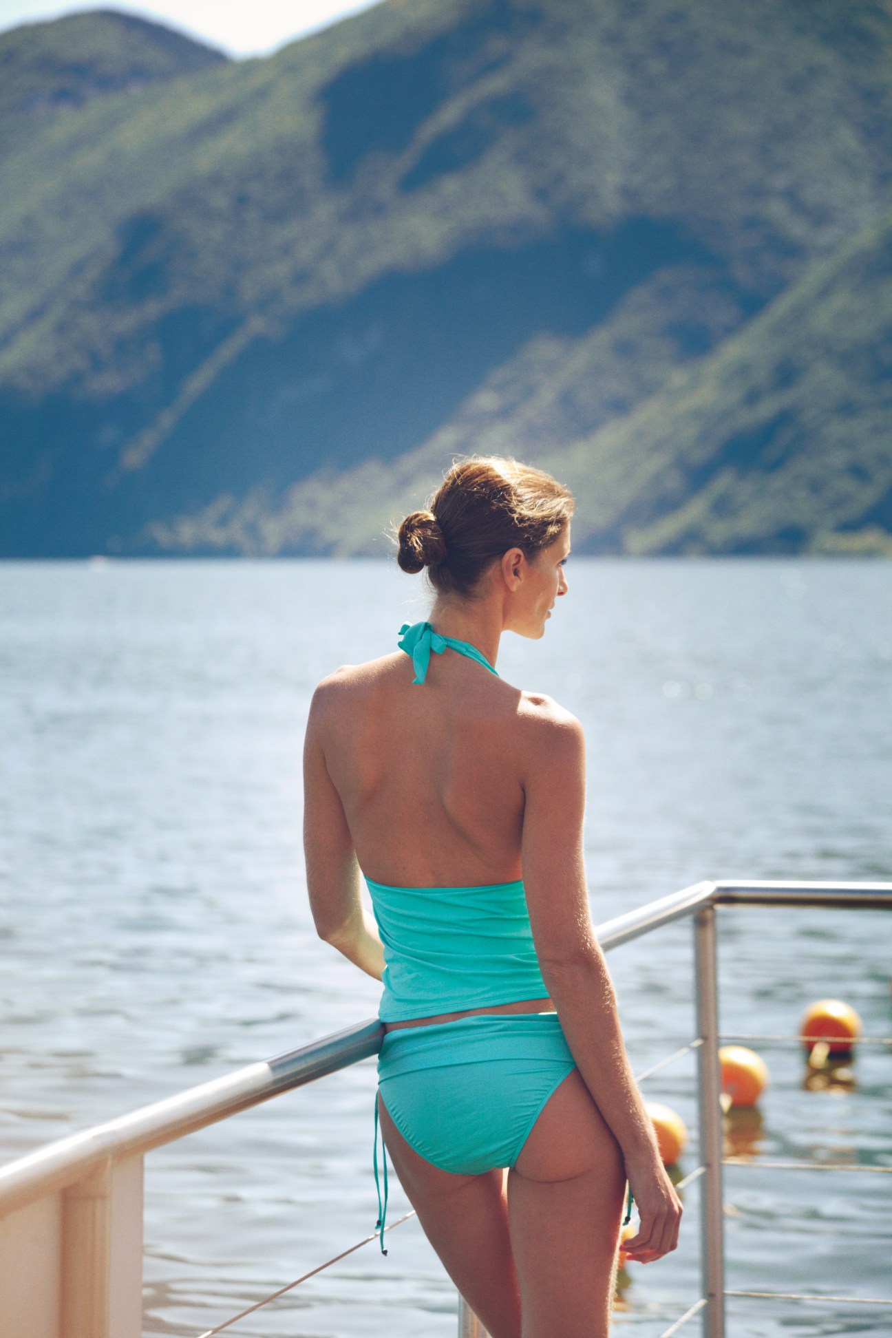 Woman wearing turquoise Polo Ralph Lauren swimwear standing by a lake with mountains in the background.