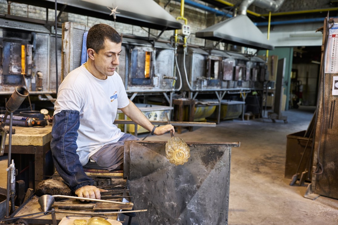 Glass-blower working with molten golden glass at a workbench in a Murano workshop.