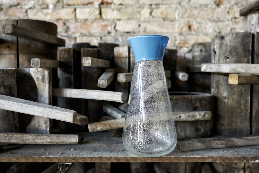 A glass vase with blue top and textured clear body displayed among wooden moulds and tools in a workshop.
