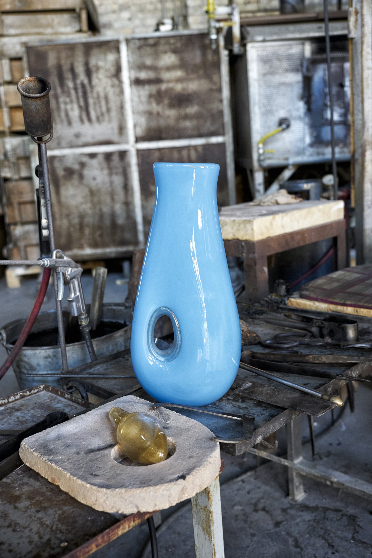 A bright blue glass vase with a narrow neck and oval opening sits on a work surface in a glass-blowing workshop, with steel tools and wooden molds visible in the background.