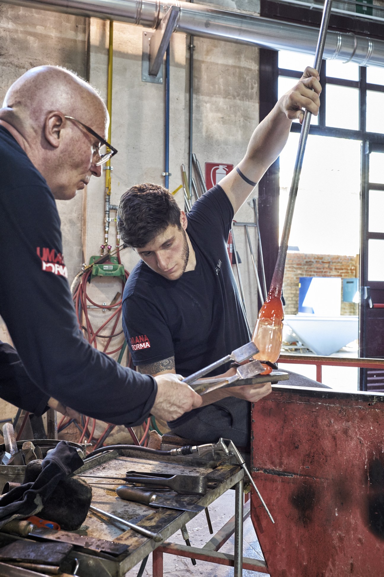 Two glass-blowers at work in a workshop, one holding a steel straw with molten glass aloft while the other observes nearby.