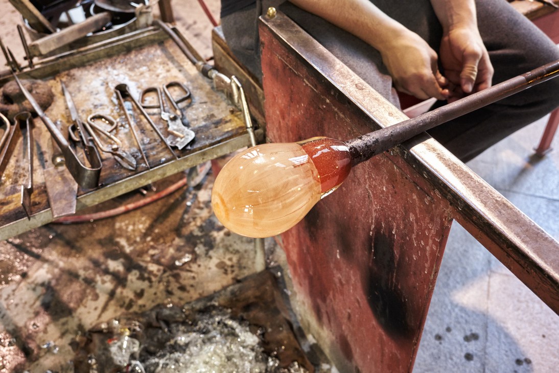 A craftsman's hands shape a molten glass bubble on a steel rod using a wooden mould without protective gloves.