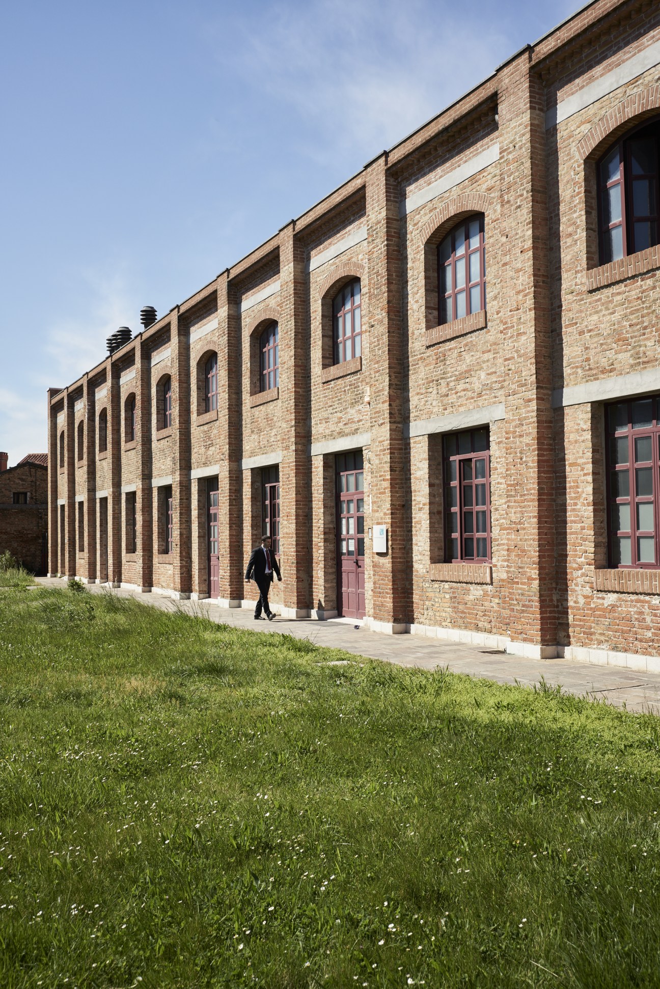 Person walking past a brick industrial building with large arched windows and red doors.