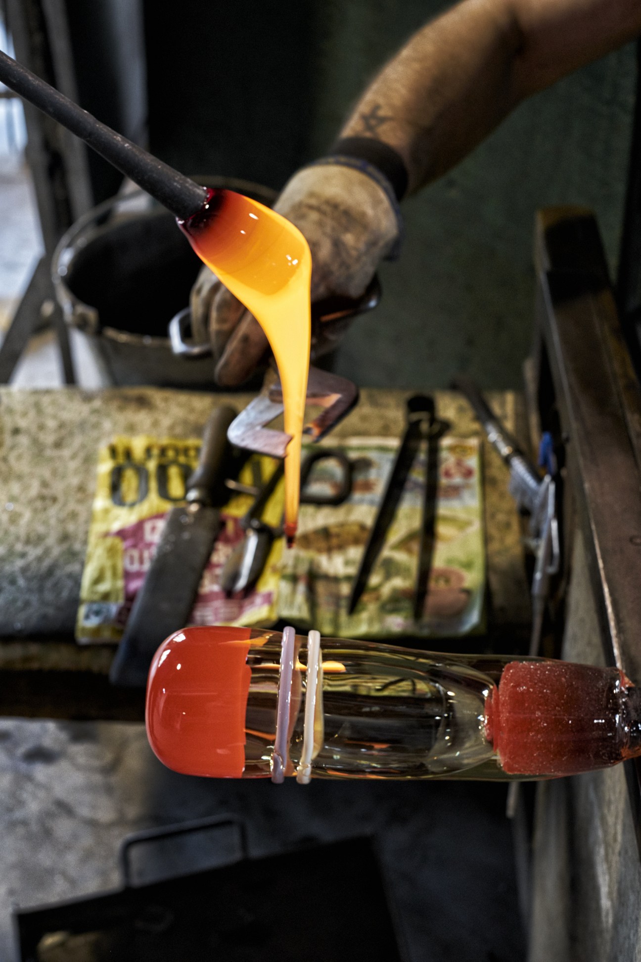A glass-blower holds a steel straw with molten yellow glass dripping onto a red glass vessel below.