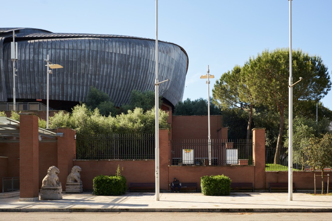 Renzo Piano's Parco della Musica with curved metallic facade, red brick wall, bronze lion sculptures, and Mediterranean pine trees in Rome.