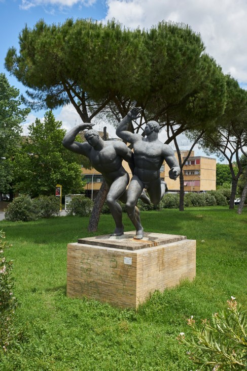 Bronze sculpture of two athletic male figures in dynamic poses under an umbrella pine tree in Rome's Olympic Village.
