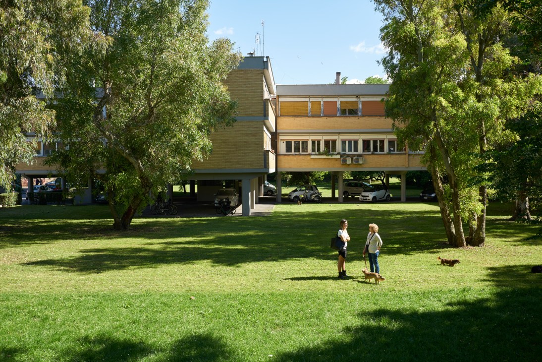 Modernist apartment building with piloti supports and residents relaxing on green lawn surrounded by trees.