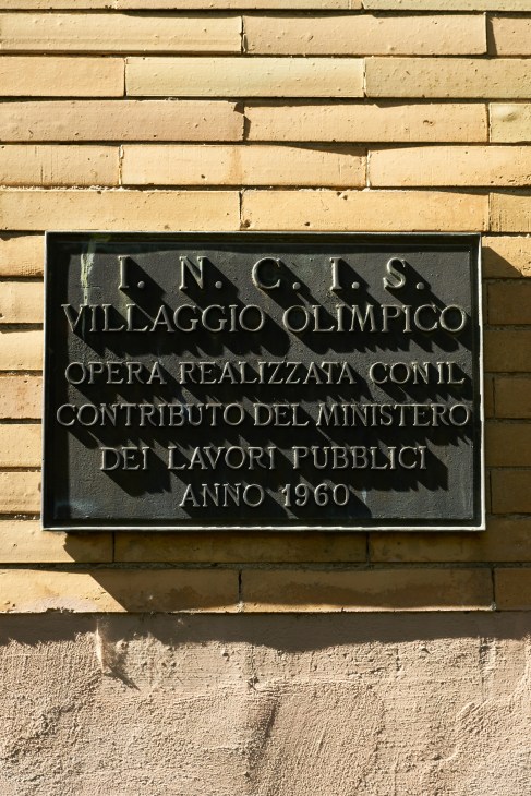 Black plaque on tan brick wall commemorating the 1960 Olympic Village construction with Italian text.