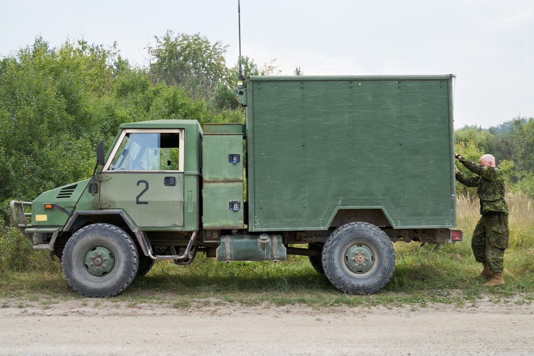 Green military truck with number 2 on door and soldier in camouflage uniform standing beside it in rural setting