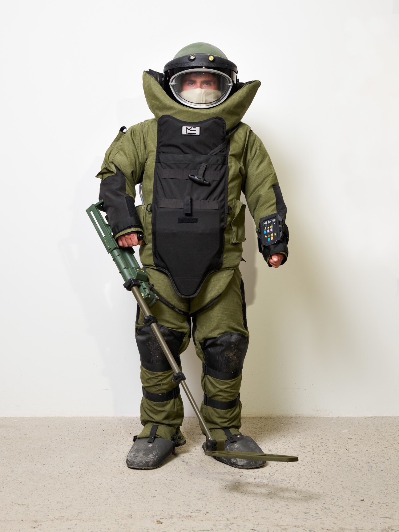 Person in olive green EOD-9 bomb disposal suit with helmet and protective vest holding detection equipment