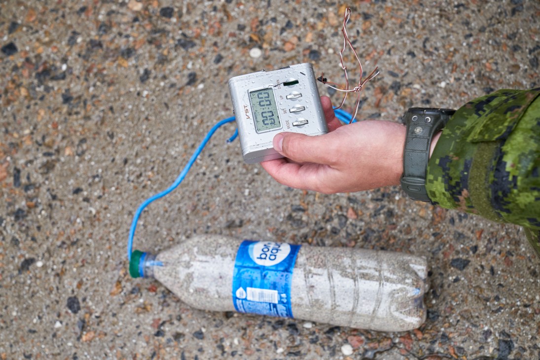 Hand holding white electronic device with digital display connected by blue cable to plastic water bottle on ground