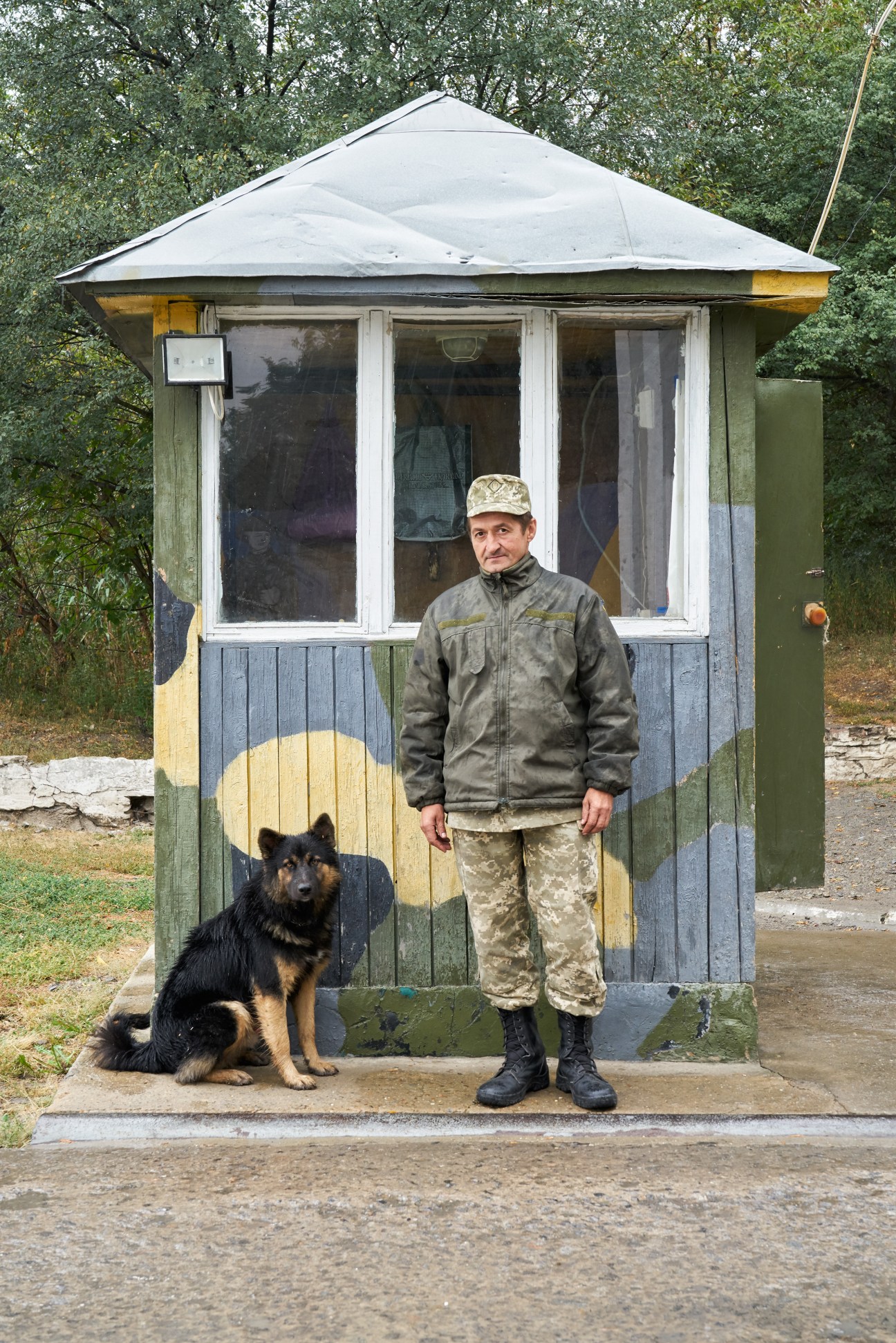 Ukrainian guard in camouflage uniform and cap stands with German Shepherd dog outside small guardhouse