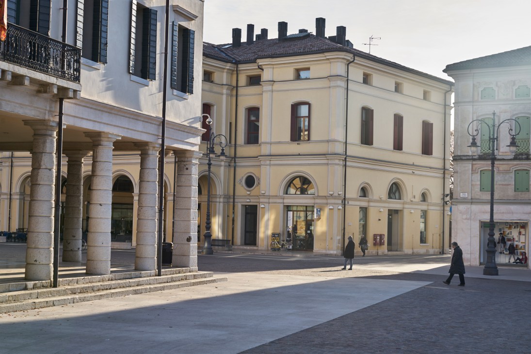 Montebelluna town square with classical Italian buildings, arcaded colonnade, and pedestrians in afternoon sunlight.