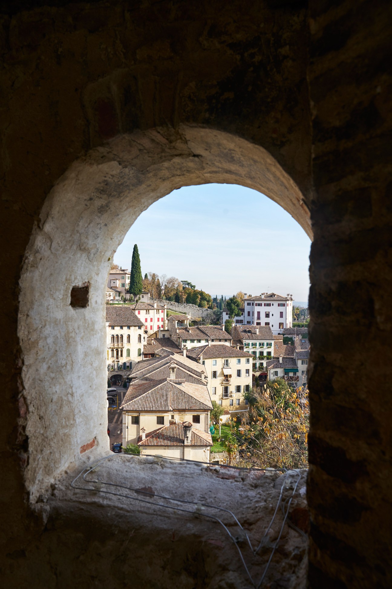 View through a stone archway of Asolo's hillside town with terracotta-roofed buildings and cypress trees.